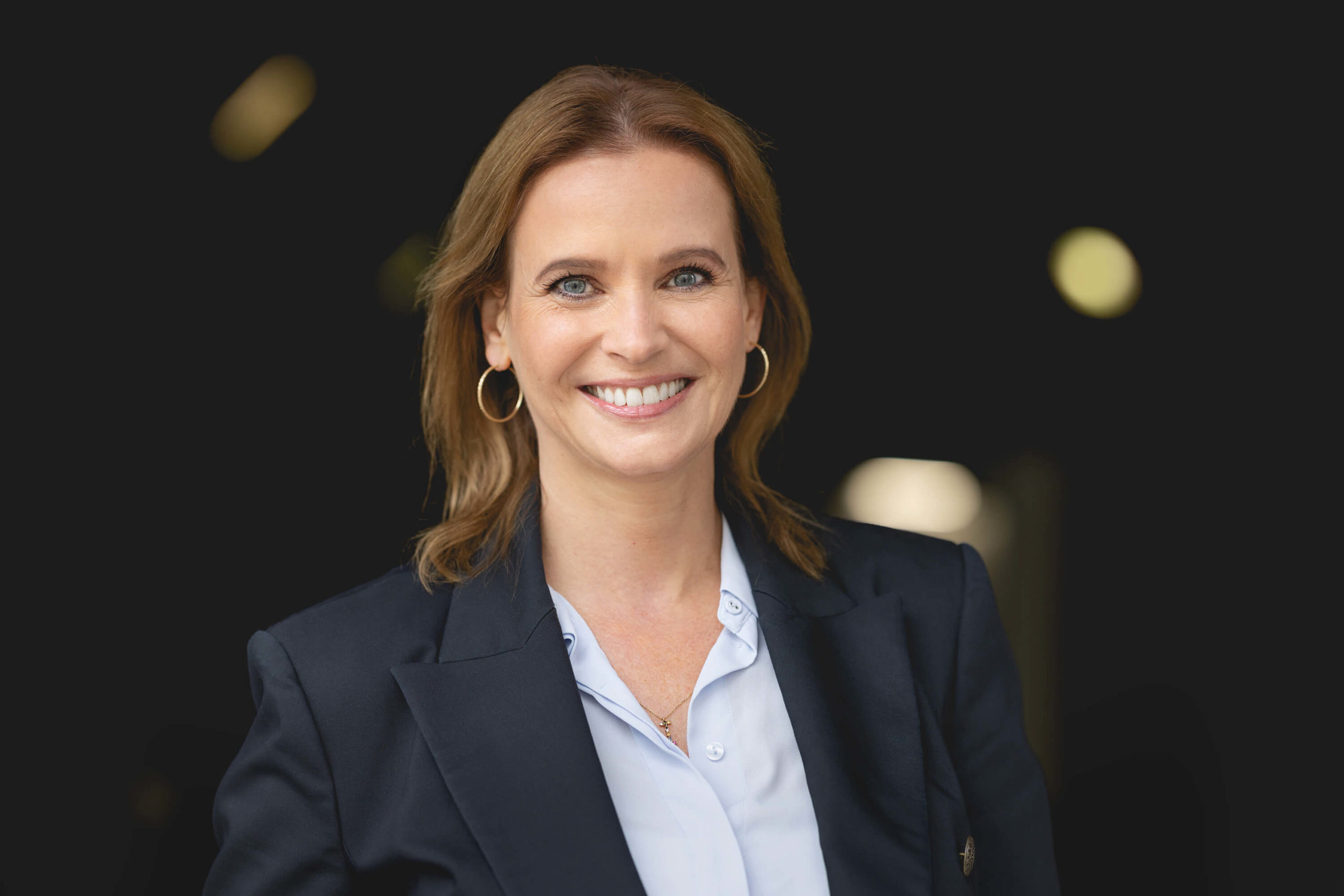 Ina Steffens HERATAX Portrait draußen Office Managerin, lächelnd blauer Blazer, hellblaue Bluse mit Kreolen, unscharfe Lichter im Hintergrund